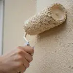 A hand rolling a fresh coat of paint onto a textured exterior wall beside a window, the kind of application that affects how long exterior paint lasts.