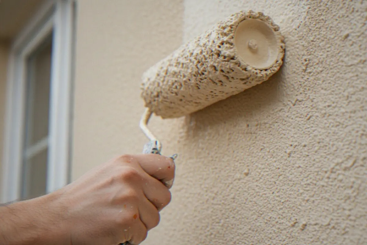 A hand rolling a fresh coat of paint onto a textured exterior wall beside a window, the kind of application that affects how long exterior paint lasts.