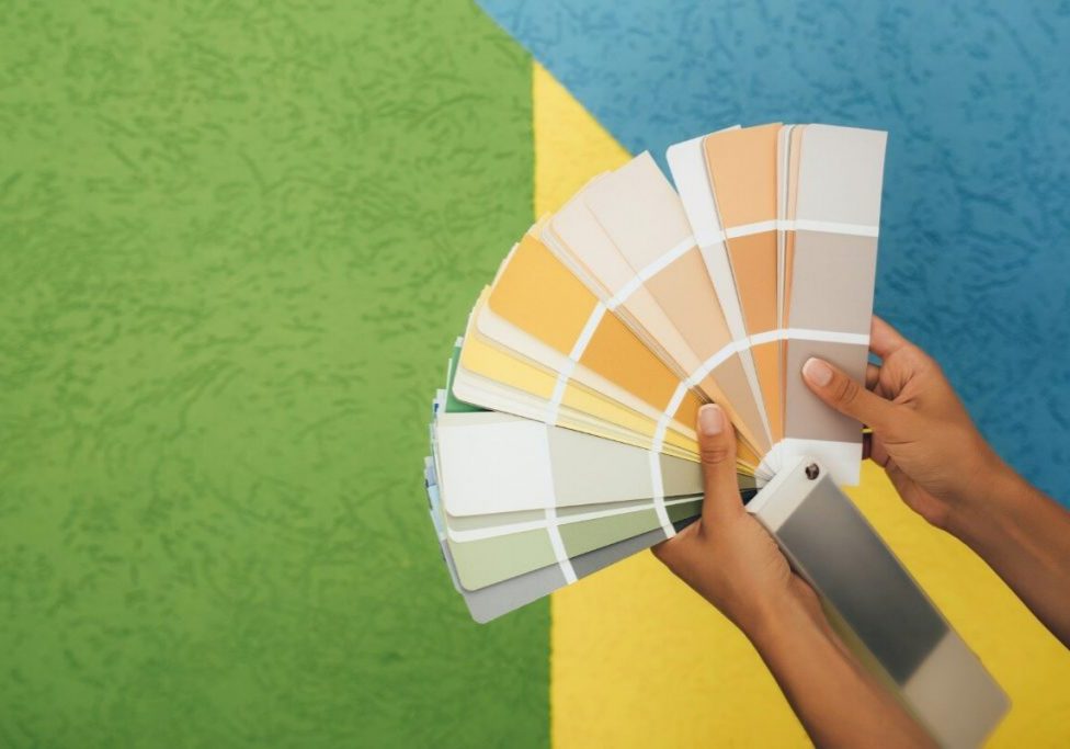 Hands holding a colorful paint swatch fan against a two-tone green and white background while learning how to choose interior paint colours.